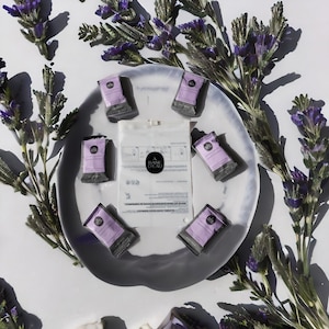 May include: A white ceramic plate with seven small, individually wrapped, purple and black foaming soap tablets. The plate is surrounded by sprigs of purple flowers.