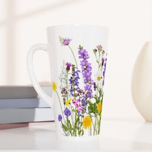 Puede incluir: Taza de cerámica blanca con un diseño alto y delgado. La taza presenta una impresión floral de flores silvestres prensadas en tonos de morado, amarillo y blanco.