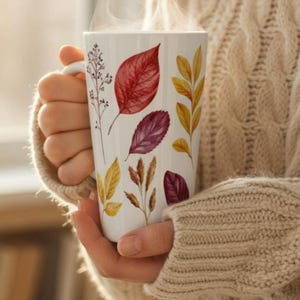 May include: White ceramic mug decorated with watercolor illustrations of autumn leaves in shades of red, yellow, and purple. The mug is held in a person's hands, and steam rises from the top, suggesting a warm beverage.