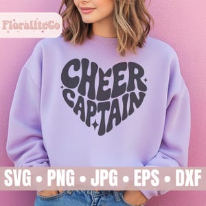 Könnte beinhalten: Ein helllila Sweatshirt mit dem Schriftzug "CHEER CAPTAIN" in Herzform, in Dunkelgrau. Das Bild zeigt auch Dateitypen: SVG, PNG, JPG, EPS und DXF. Der Hintergrund ist rosa.