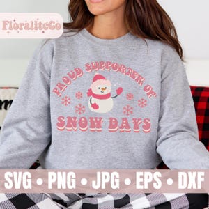 May include: A gray sweatshirt with a pink graphic that reads "Proud Supporter of Snow Days." The graphic features a snowman wearing a pink hat and scarf, surrounded by snowflakes. The brand name "FloraliteCo" is in the top left corner.