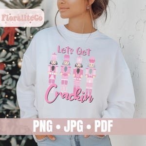 Peut inclure: Un sweat-shirt blanc à manches longues avec une impression graphique de quatre casse-noisettes roses alignés. Au-dessus des casse-noisettes, le texte indique "Lets Get" et en dessous, le texte "Crackin" en police rose grasse.