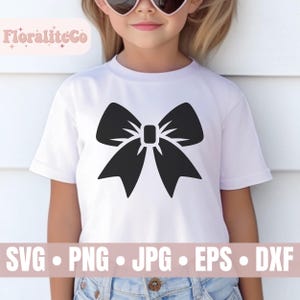 Op de afbeelding: Een wit T-shirt met een zwart strikdesign. Het shirt wordt gedragen door een kind. De tekst "FloraliteCo" is zichtbaar in de linkerbovenhoek van de afbeelding.