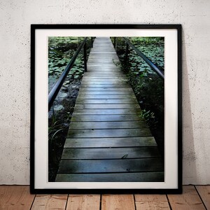 May include: A framed photograph showing a wooden boardwalk over a pond with lily pads. The boardwalk has dark metal railings. The image is in a black frame, set against a textured white wall. The floor is wooden.