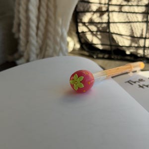 Peut inclure: Un stylo rose avec un motif de fraise sur le dessus, avec une fleur verte et des points jaunes. Le stylo repose sur un cahier ouvert avec des pages blanches vierges. Le corps du stylo est orange.
