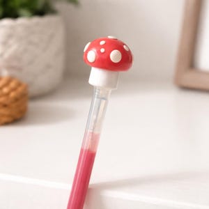 May include: A red mushroom-topped pen with white polka dots. The pen has a clear barrel filled with pink ink. The pen is a whimsical stationery item.