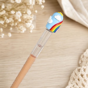 May include: A clear pen with a rainbow and cloud design on the top. The pen's tip is a light peach color. The pen is on a light wood surface.