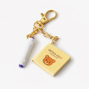 May include: A gold-colored keychain with a lobster clasp, chain, and two charms. One charm is a white marker with a purple tip. The other is a small yellow book with a brown bear illustration and the text "CUTE & COZY COLORING BOOK."