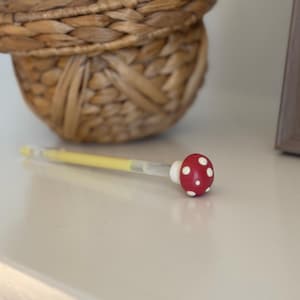 以下が含まれることがあります： 赤いキノコ型のキャップに白い水玉模様が付いた装飾的なペン。ペンの軸は黄色で、先端は透明です。白い表面に置かれており、背景には編まれたバスケットがあります。ペンの長さは約15cmです。