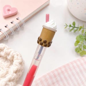May include: A novelty pen designed to resemble a bubble tea drink. The pen features a brown cup with dark brown boba pearls, a white whipped cream topping, and a pink straw. The pen's barrel is clear with red ink.