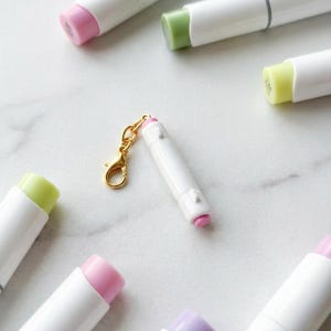 May include: A white lip balm charm with a pink cap and gold-colored clasp. The charm is surrounded by several white lip balm tubes with various colored caps, including pink, green, and yellow. The background is a light marble surface.