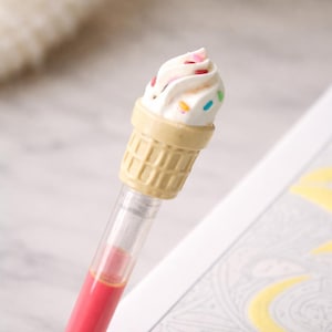 May include: A novelty pen featuring an ice cream cone design. The pen has a clear barrel filled with red ink. The ice cream cone is beige with a white ice cream top, decorated with colorful sprinkles.