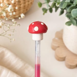 May include: A red mushroom-shaped pen with white polka dots. The pen has a white base and a clear barrel filled with pink ink. The pen is a whimsical and decorative writing tool.