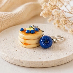 Könnte beinhalten: Ein Miniatur-Schlüsselanhänger mit gestapelten Pfannkuchen, belegt mit Blaubeeren und einem Butterstück. Ein großer blauer Blaubeer-Anhänger ist befestigt. Die Pfannkuchen sind mit Sirup glasiert. Der Schlüsselanhänger ist aus Polymer Clay.