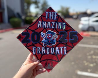 May include: A red graduation cap with a spiderweb design and the words "THE AMAZING GRADUATE" in white and blue. The numbers "2025" are in black and blue, with a Spider-Man graphic.