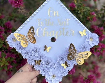 May include: A light blue graduation cap decorated with white paper flowers and gold butterflies. The cap has a gold inscription that reads "On to the Next Chapter".