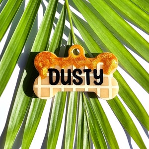 Pode incluir: Uma etiqueta para cachorro em forma de osso com um padrão de waffle e detalhes de glitter dourado. A etiqueta tem o nome "DUSTY" escrito em letras pretas.