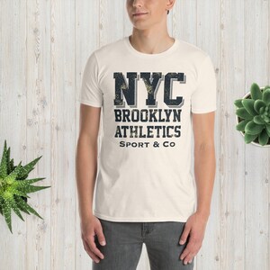 May include: A white t-shirt with a black and gray graphic print that says "NYC Brooklyn Athletics Sport & Co."