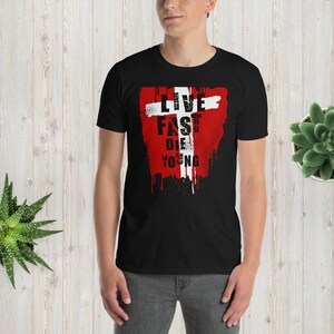 Op de afbeelding: Zwart T-shirt met een rood en wit grafisch ontwerp. De grafiek toont een wit kruis met de woorden "Live Fast Die Young" erop.
