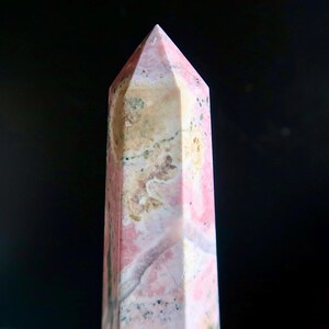 May include: A pink and beige gemstone obelisk with a pointed top. The stone has a natural, mottled pattern with variations in colour and texture. The obelisk is set against a dark background.