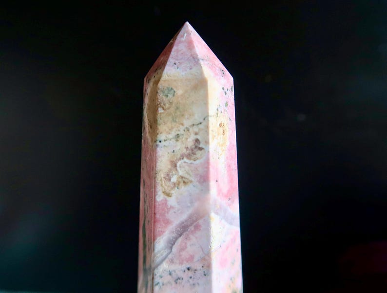 May include: A pink and beige gemstone obelisk with a pointed top. The stone has a natural, mottled pattern with variations in colour and texture. The obelisk is set against a dark background.