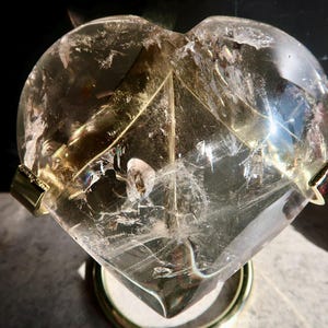 Puede incluir: Un cristal de cuarzo transparente en forma de corazón con fracturas e inclusiones internas, que muestra un tono dorado pálido. La piedra está sostenida por un soporte de metal dorado con dos clips laterales y una base circular, reflejando la luz.