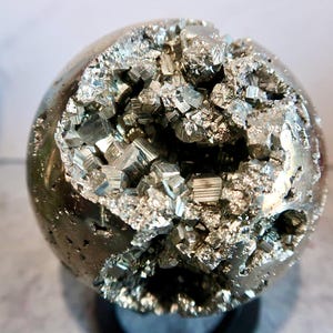 Könnte beinhalten: Eine polierte, kugelförmige Pyrit-Geode. Das Äußere hat einen metallischen Glanz, während das Innere eine Ansammlung von funkelnden, kubischen Pyritkristallen aufweist. Die Oberfläche der Geode ist eine Mischung aus glatten und rauen Texturen, die natürliche Formationen zeigen.