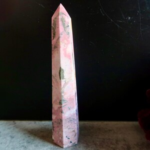 May include: A tall, pink and white obelisk-shaped crystal with a pointed top. The crystal has a marbled appearance with streaks of darker colours. The crystal is standing upright on a grey surface against a dark background.