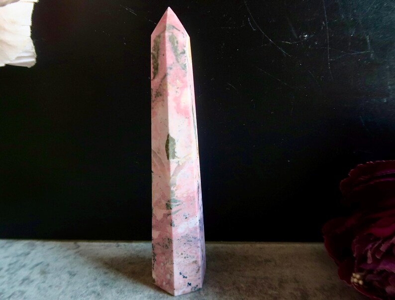 May include: A tall, pink and white obelisk-shaped crystal with a pointed top. The crystal has a marbled appearance with streaks of darker colours. The crystal is standing upright on a grey surface against a dark background.