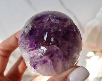 May include: A spherical amethyst crystal geode with a deep purple hue. The crystal has a rough, textured surface with visible crystalline structures and a lighter, almost white, base. The geode is held in a hand.