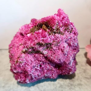 May include: A raw, crystalline mineral specimen featuring a vibrant, saturated magenta-pink color. The surface is highly textured with numerous small, jagged crystal points and clusters, interspersed with patches of dark brown, earthy mineral deposits.