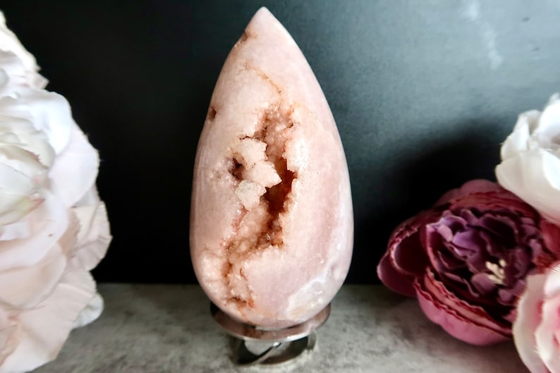 Puede incluir: Una geoda rosa en forma de l&aacute;grima con un interior forrado de cristales. La geoda est&aacute; sobre un soporte plateado.