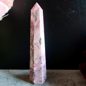 May include: A pink and grey obelisk-shaped crystal with a pointed top. The crystal has a polished surface and a mix of pink and grey tones. The crystal is standing upright against a dark background.