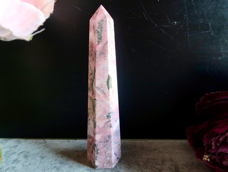 May include: A pink and grey obelisk-shaped crystal with a pointed top. The crystal has a polished surface and a mix of pink and grey tones. The crystal is standing upright against a dark background.