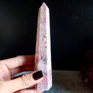 May include: A tall, pink and white crystal obelisk with a pointed top. The obelisk has a rectangular shape with angled sides. The crystal has a marbled appearance with dark grey and black inclusions. The obelisk is held in a hand.