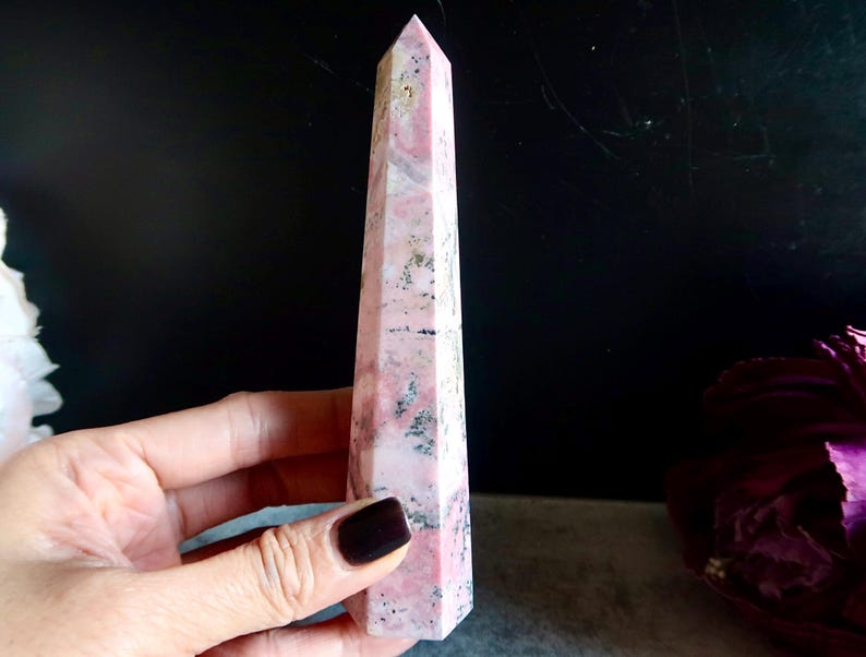 May include: A tall, pink and white crystal obelisk with a pointed top. The obelisk has a rectangular shape with angled sides. The crystal has a marbled appearance with dark grey and black inclusions. The obelisk is held in a hand.