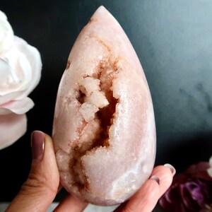 Puede incluir: Un cristal rosa en forma de l&aacute;grima con una gran cavidad abierta llena de cristales m&aacute;s peque&ntilde;os. El cristal es liso y pulido, con un tono rosa claro.