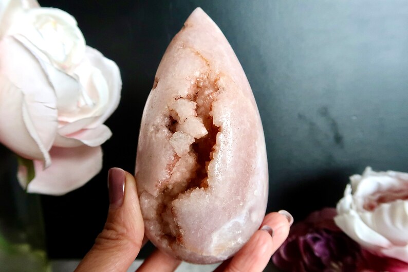 Puede incluir: Un cristal rosa en forma de l&aacute;grima con una gran cavidad abierta llena de cristales m&aacute;s peque&ntilde;os. El cristal es liso y pulido, con un tono rosa claro.