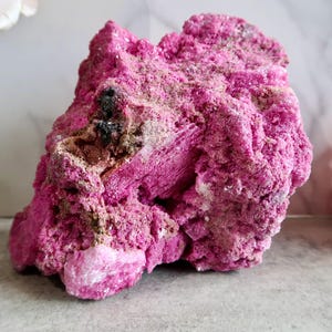Peut inclure: Un spécimen minéral brut d'un magenta vibrant avec une texture cristalline rugueuse. La pierre présente des amas rose foncé, de petites taches de dépôts minéraux blancs et une inclusion brun-noir sombre incrustée dans la surface irrégulière.