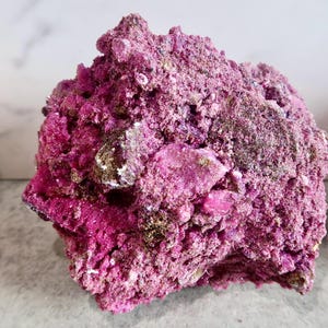May include: A raw, jagged mineral specimen featuring a vibrant magenta and deep pink coloration. The surface is highly textured with crystalline formations, irregular crevices, and small patches of dark brown and metallic gold mineral deposits.