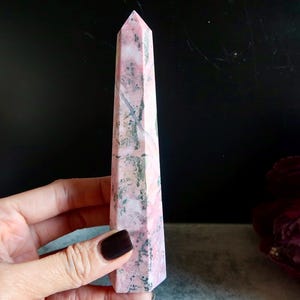 May include: A pink and grey gemstone obelisk, held in a hand. The obelisk is a tall, four-sided structure with a pointed top. The stone has a marbled appearance with varying shades of pink and grey. The obelisk is a decorative item.