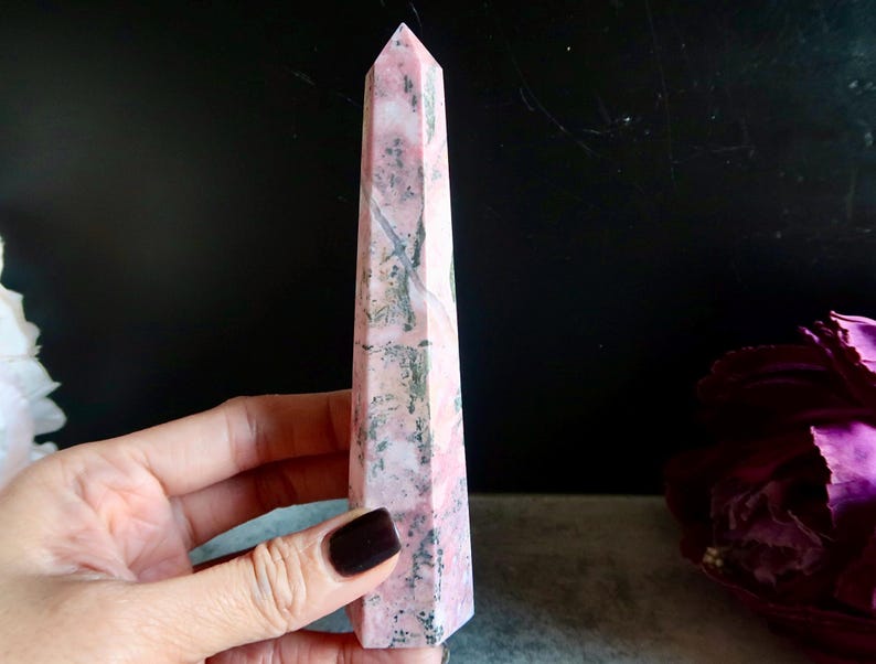 May include: A pink and grey gemstone obelisk, held in a hand. The obelisk is a tall, four-sided structure with a pointed top. The stone has a marbled appearance with varying shades of pink and grey. The obelisk is a decorative item.
