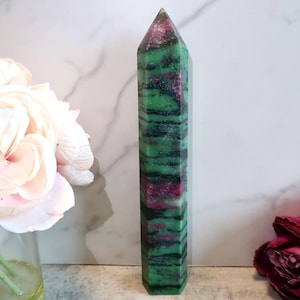 Könnte beinhalten: Ein hoher, rechteckiger Rubin-Zoisit-Kristall-Obelisk. Der polierte Stein weist Bänder aus Grün, Schwarz und Rosa auf. Der Obelisk steht aufrecht, mit einer spitzen Spitze. Blumen sind im Hintergrund.