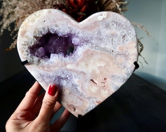 May include: A heart-shaped amethyst geode with a light pink and white exterior. The geode has a cavity filled with sparkling purple crystals. The geode is displayed on a black stand, showcasing its natural beauty.