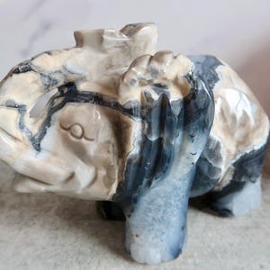 May include: A carved stone elephant figurine in shades of cream, gray, and black. The elephant is depicted with its trunk raised and its head tilted. The figurine is approximately 4 inches long and 3 inches tall.