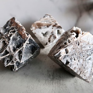May include: Three square-shaped, natural stone specimens with intricate, porous patterns. The stones display a mix of grey, white, and brown hues, creating a textured appearance. The objects are arranged on a light-coloured surface.
