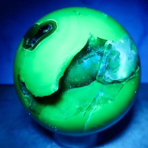 May include: A spherical mineral specimen glowing with a bright neon green fluorescence under ultraviolet light. The surface features smooth, polished areas contrasted by irregular, recessed crystalline cavities and natural fissures.