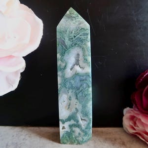 May include: A polished moss agate crystal tower featuring a pointed top and a hexagonal base. The stone displays a complex pattern of deep forest green, translucent white, and grey inclusions with small, naturally occurring geode cavities.