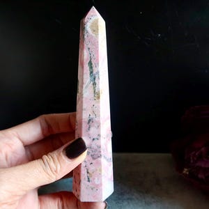 May include: A pink and beige crystal obelisk with a pointed top. The stone has a marbled appearance with dark grey and white veining. The obelisk is held in a hand against a dark background.