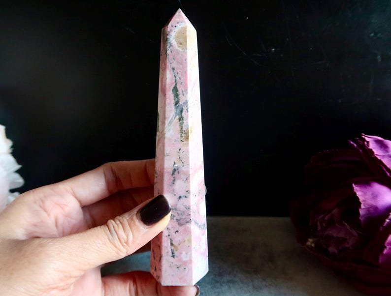 May include: A pink and beige crystal obelisk with a pointed top. The stone has a marbled appearance with dark grey and white veining. The obelisk is held in a hand against a dark background.
